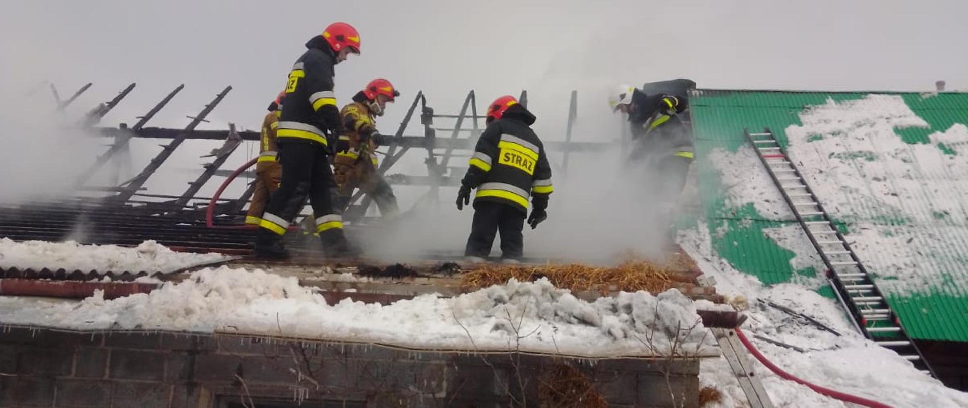Na zdjęciu widoczny budynek ze spalonym dachem. Strażacy gaszący pożar na poddaszu. Z prawej strony widoczny zielony dach o który oparte są drabiny pożarnicze.