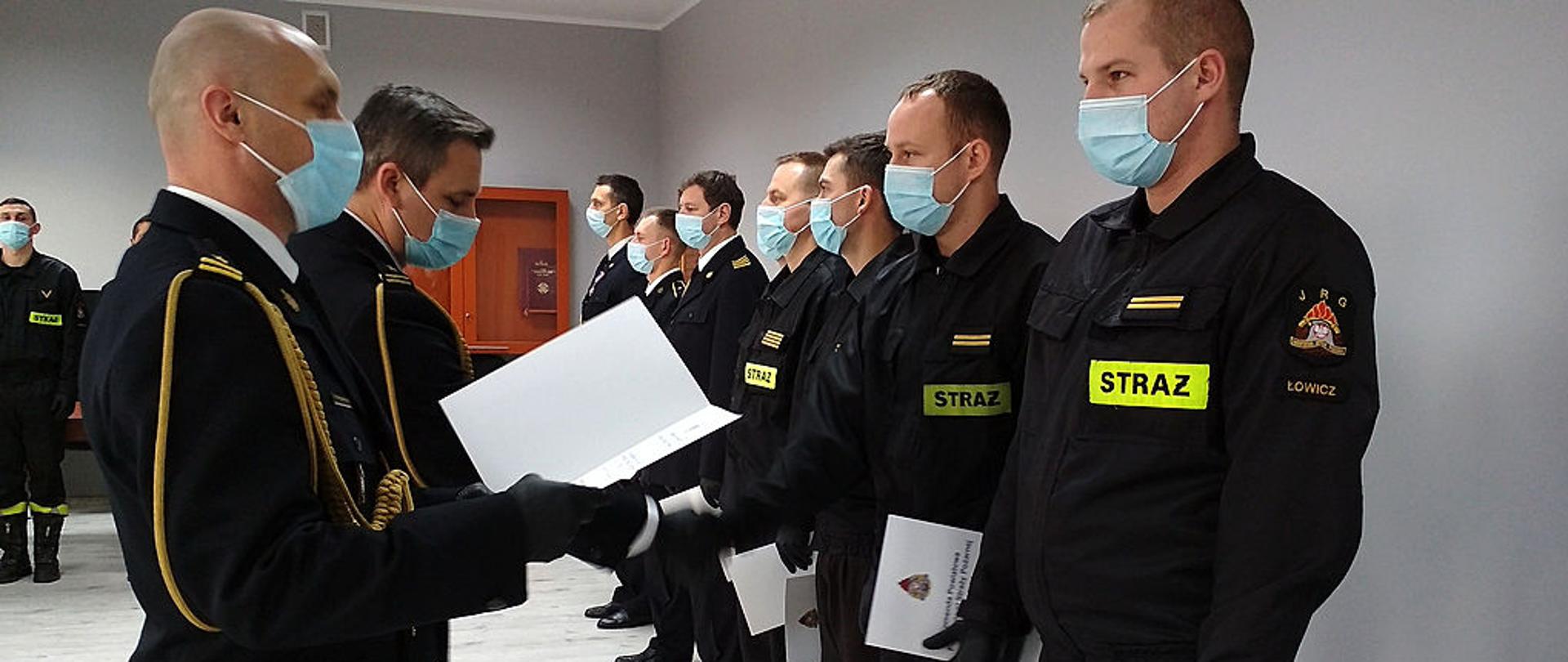 na zdjęciu komendant powiatowy PSP w Łowiczu mł. bryg. Arkadiusz Makowski oraz zastępca komendanta powiatowego mł. bryg. Rafał Garbarz wręczają odznaczenia i awanse w stopniach dla funkcjonariuszy KP PSP w Łowiczu którzy stoją w szeregu w pomieszczeniu sali konferencyjnej. Strażacy ubrani są w mundury galowe ze sznurem oraz ubrania koszarowe.