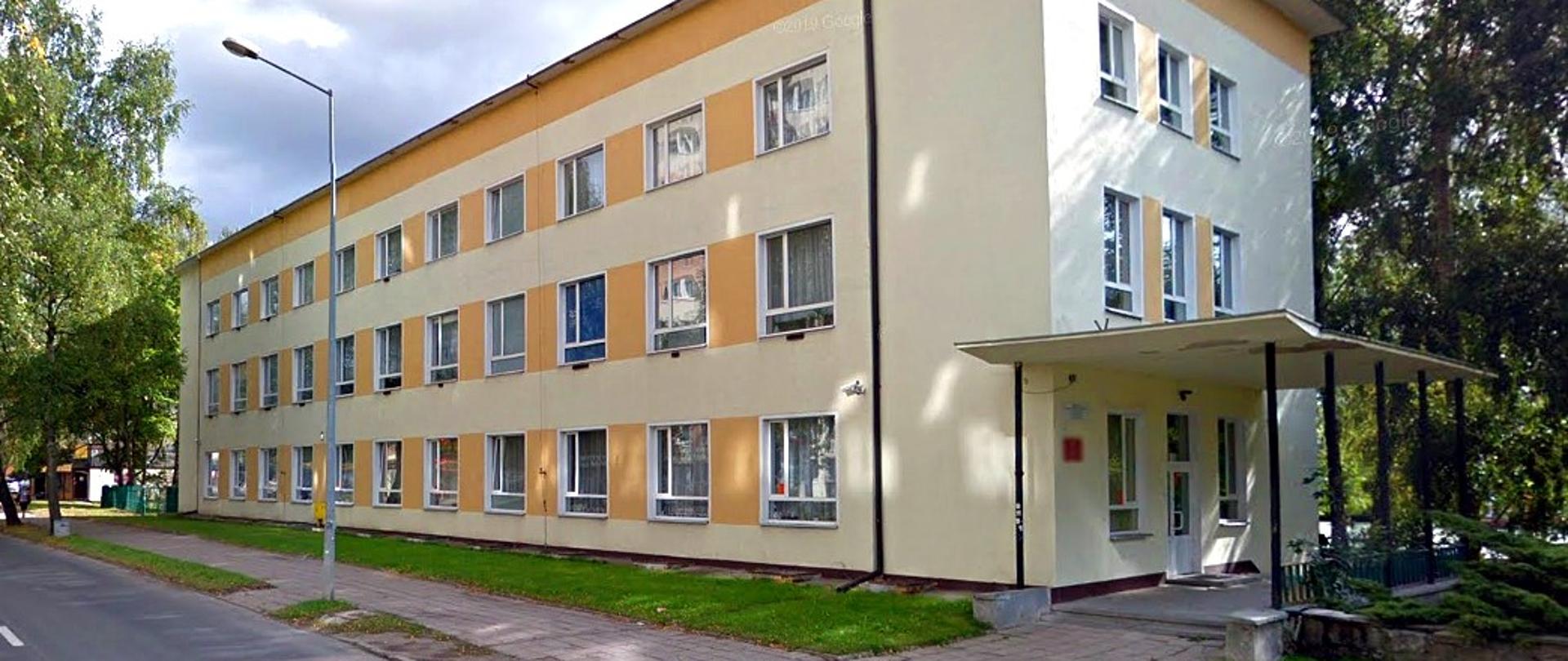 z lewej strony zdjęcia fragmentu jezdni i chodnika, po prawej trzykondygnacyjny budynek z równomiernie rozłożonymi oknami wzdłuż chodnika, od prawej wejście główne z daszkiem wspartym na wąskich kolumnach