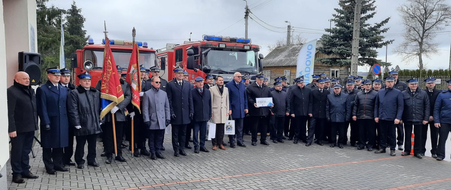 Zdjęcie grupowe uczestników uroczystości na tle samochodów pożarniczych. 
