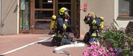 Na zdjęciu widzimy strażaków ubierających sprzęt ochrony dróg oddechowych na tle budynku, w którym przeprowadzono ćwiczenia.