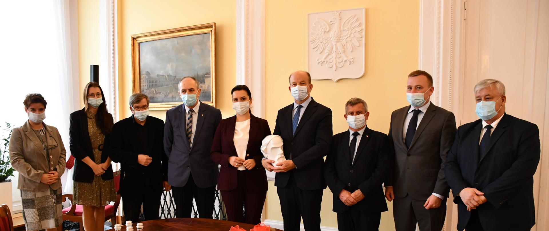 Na zdjęciu znajduje się 9 elegancko ubranych osób - 3 kobiety i 6 mężczyzn. Osoby te pozują na tle Godła RP, umieszczonego na fragmencie ściany w kolorze jasnożółtym. Tuż przed osobami znajduje się fragment owalnego, drewnianego stołu. 