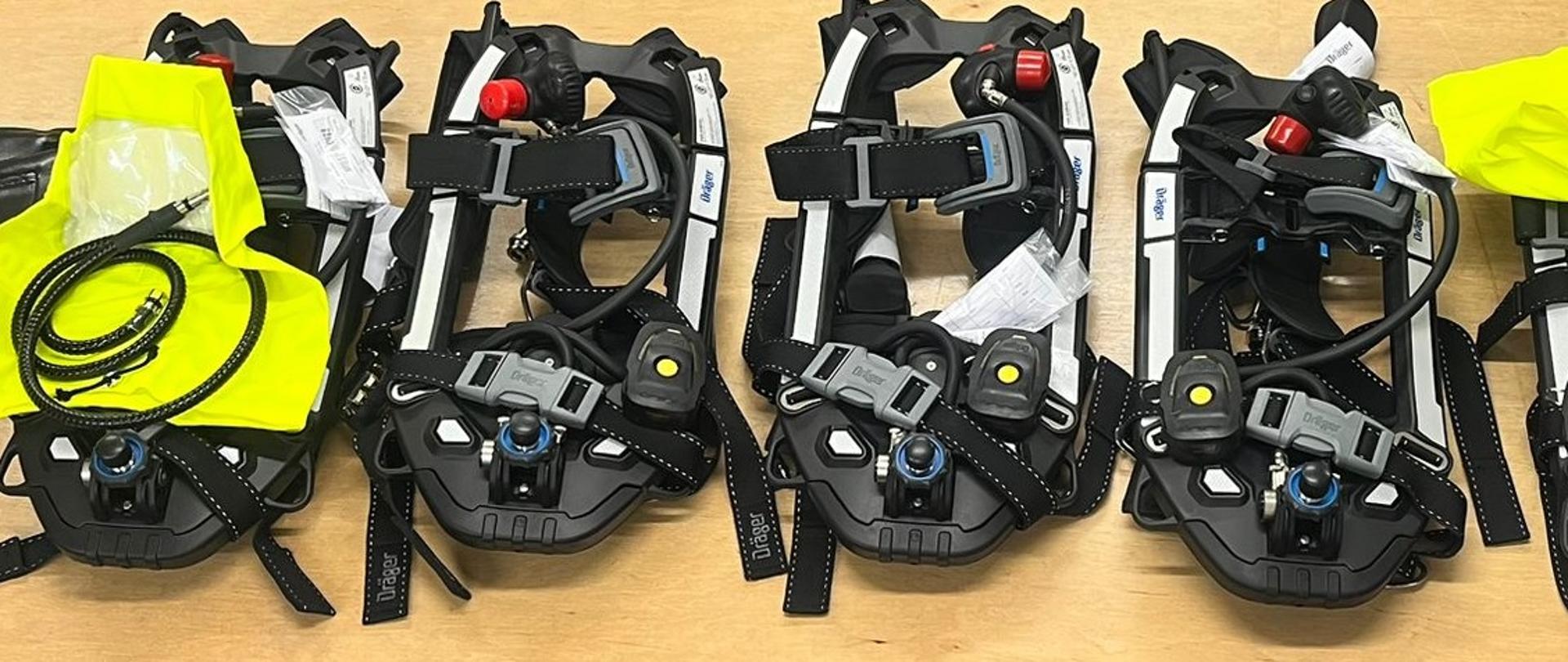 Zdjęcie przedstawia 4 aparaty powietrzne koloru czarnego z białymi paskami wraz z 2 kapturami ucieczkowymi koloru jaskrawo żółtego leżące na drewnianym stole.