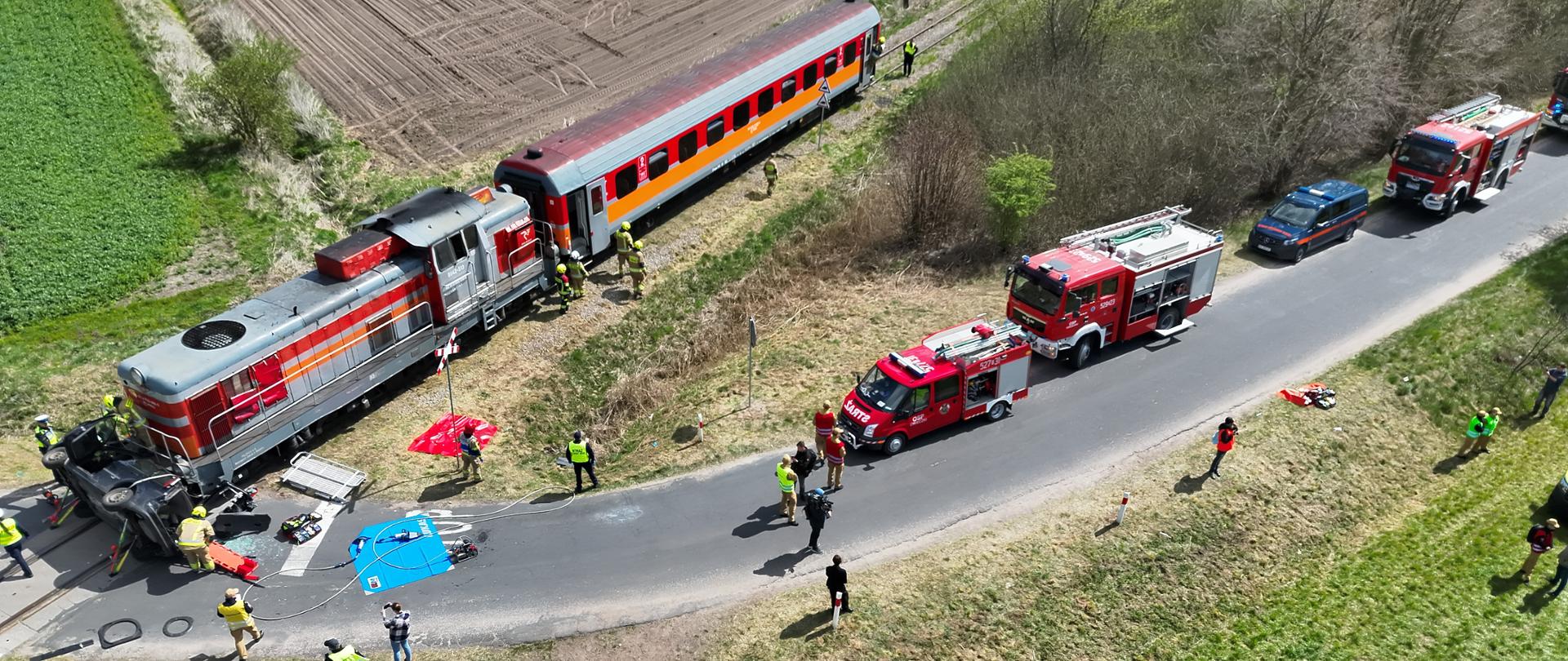 Zdjęcie, wykonane z góry, widać na nim miejsce wypadku na przejeździe kolejowym: pociąg zatrzymał się przy uszkodzonym samochodzie, a wokół pracują strażacy i służby ratunkowe.