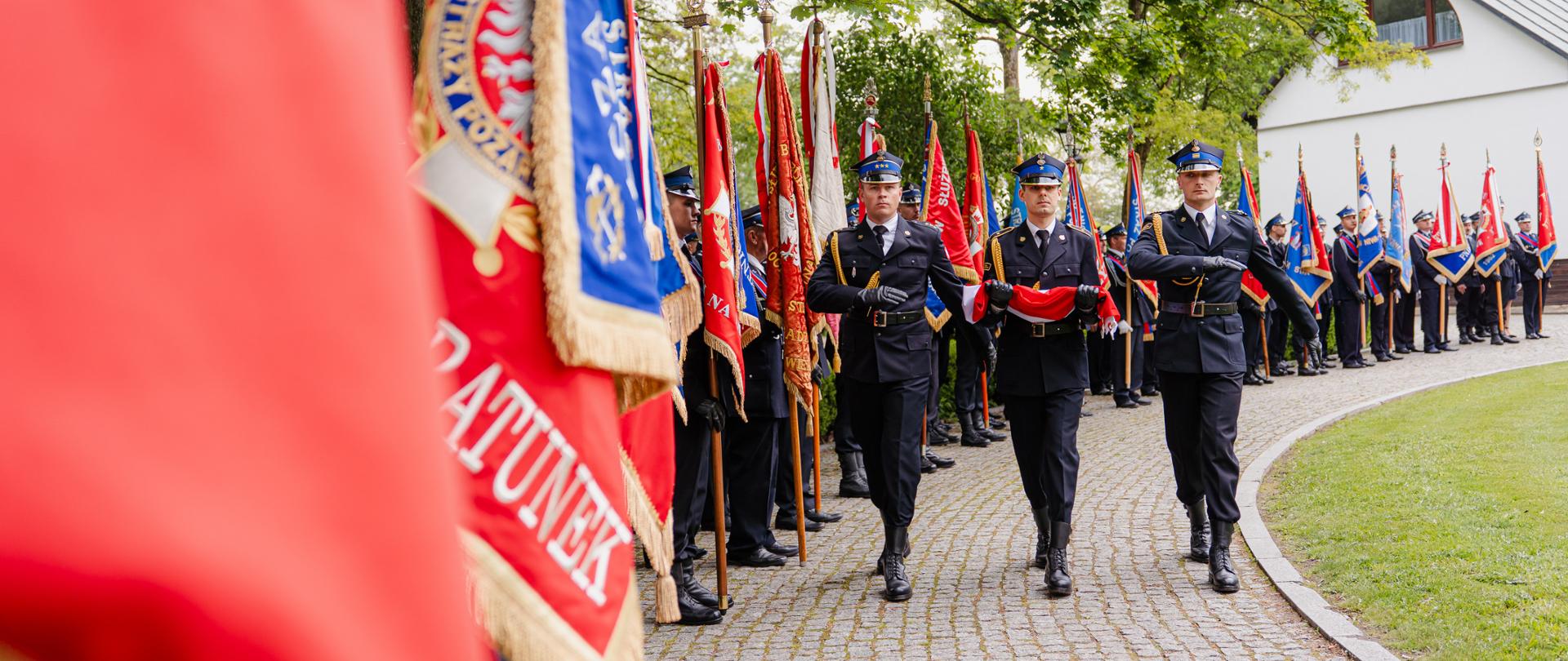 Dzień Strażaka 2025 w powiecie siedleckim. Trzech strażaków maszeruje po chodniku obok długiego pododdziału strażackich pocztów sztandarowych. 