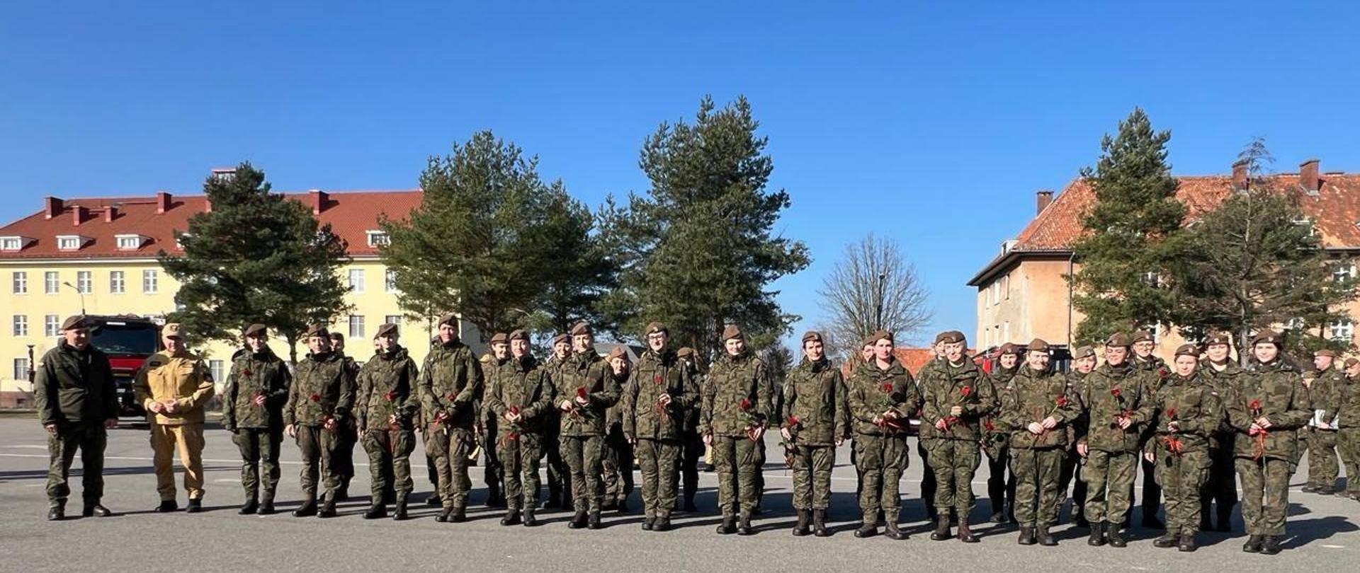 Na zdjęciu duża grupa żołnierzy WOT, kobiet. Trzymają w rekach czerwone róże. Niebieskie niebo,słonecznie. Z lewej strony dowódca WOT i komendant powiatowy PSP.