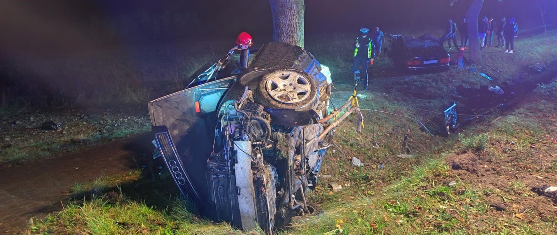 Zdjęcie przedstawia miejsce wypadku przy drodze wojewódzkiej. W centralnym punkcie znajduje się część rozerwanego samochodu osobowego, przy nim stoi jeden strażak. Za nim rośnie drzewo w które uderzyło auto. W głębi po prawej stronie widać drugą część pojazdu oraz funkcjonariusza Policji.