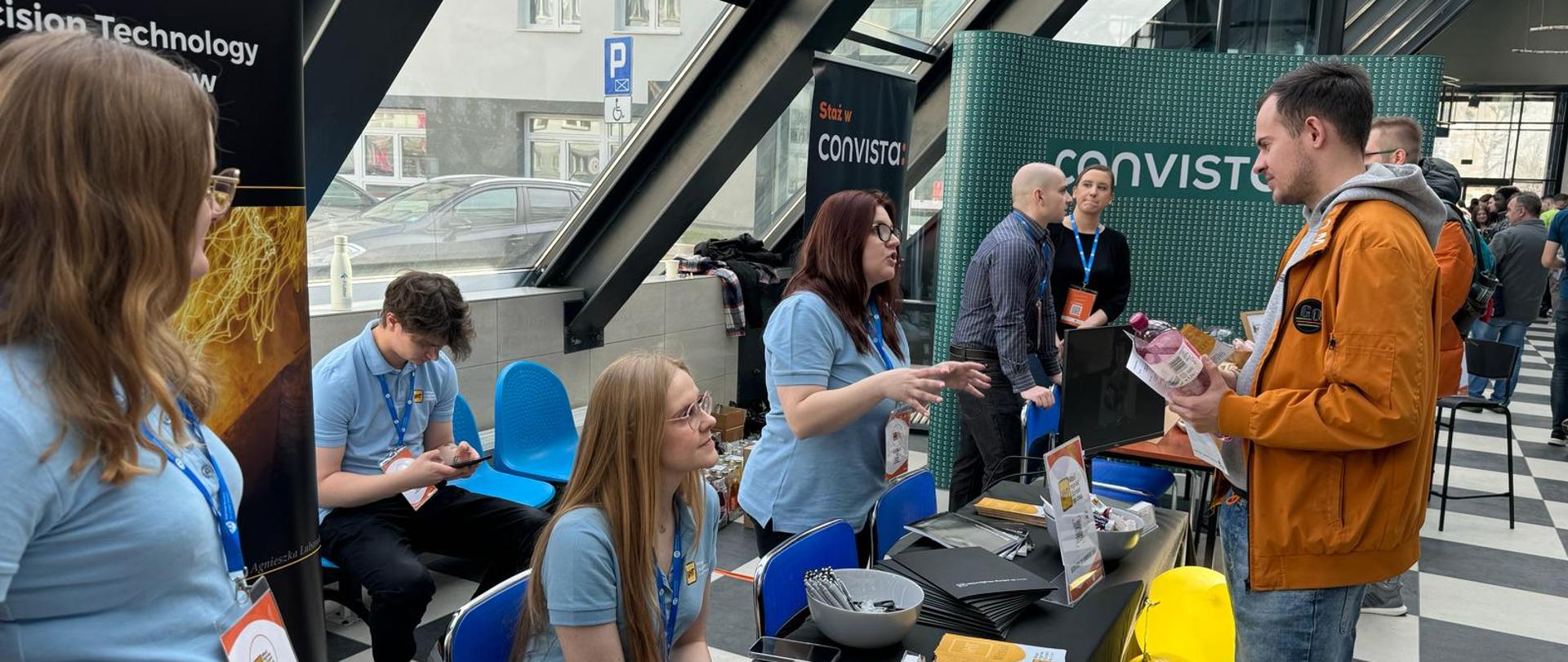 Jedno ze stanowisk dostępnych dla odwiedzających na XV Akademickich Targach Pracy Politechniki Opolskiej. Na zdjęciu widać osoby ze stanowiska, które wszystkie są ubrane w niebieskie koszulki. Jedna z tych osób rozmawia z odwiedzającym targi. Uczestnik ten ubrany jest w pomarańczową kurtkę. Słucha tego, co mówi do niego pani ze stanowiska. Na zdjęciu widać również stół, na którym znajdują się darmowe gadżety, ulotki i informacje dotyczące danego pracodawcy. 