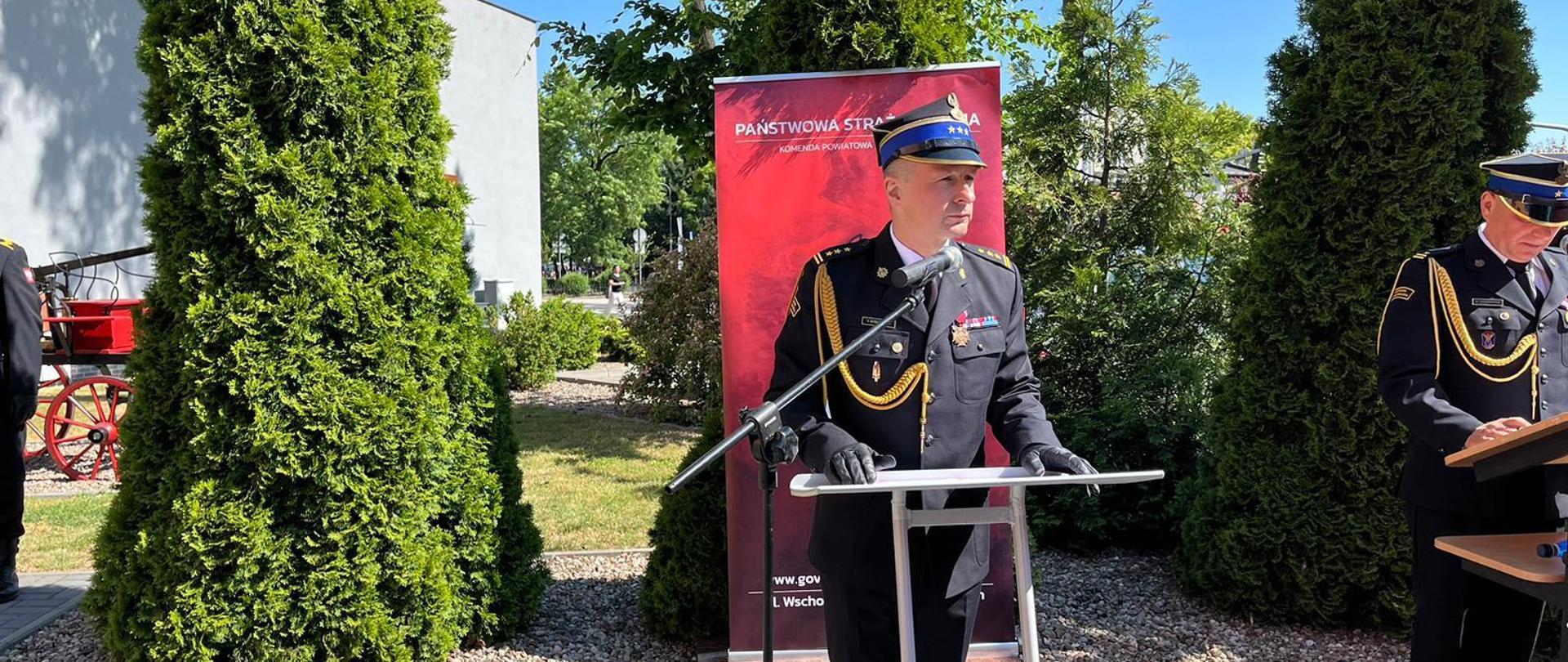 przemówienie zastępcy wielkopolskiego komendanta wojewódzkiego PSP , w tle zielone drzewa i baner