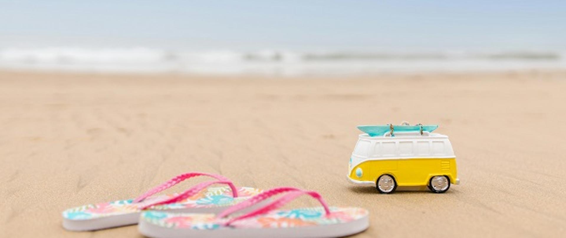 na zdjęciu widać leżące na piasku na plaży klapki-japonki oraz zabawkę samochód, w tle na drugim planie akwen przypominający jezioro lub morze