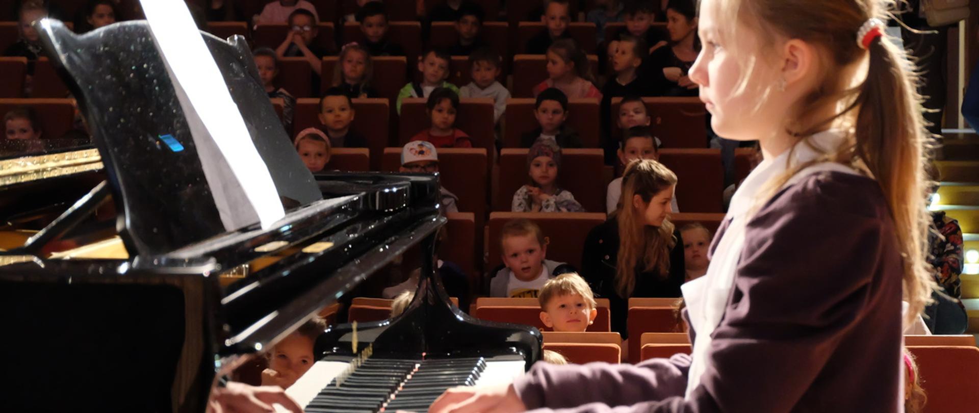Dziewczynka gra na fortepianie. W tle na widowni dużo dzieci.