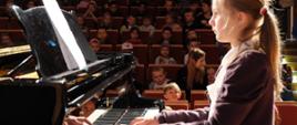 Dziewczynka gra na fortepianie. W tle na widowni dużo dzieci.