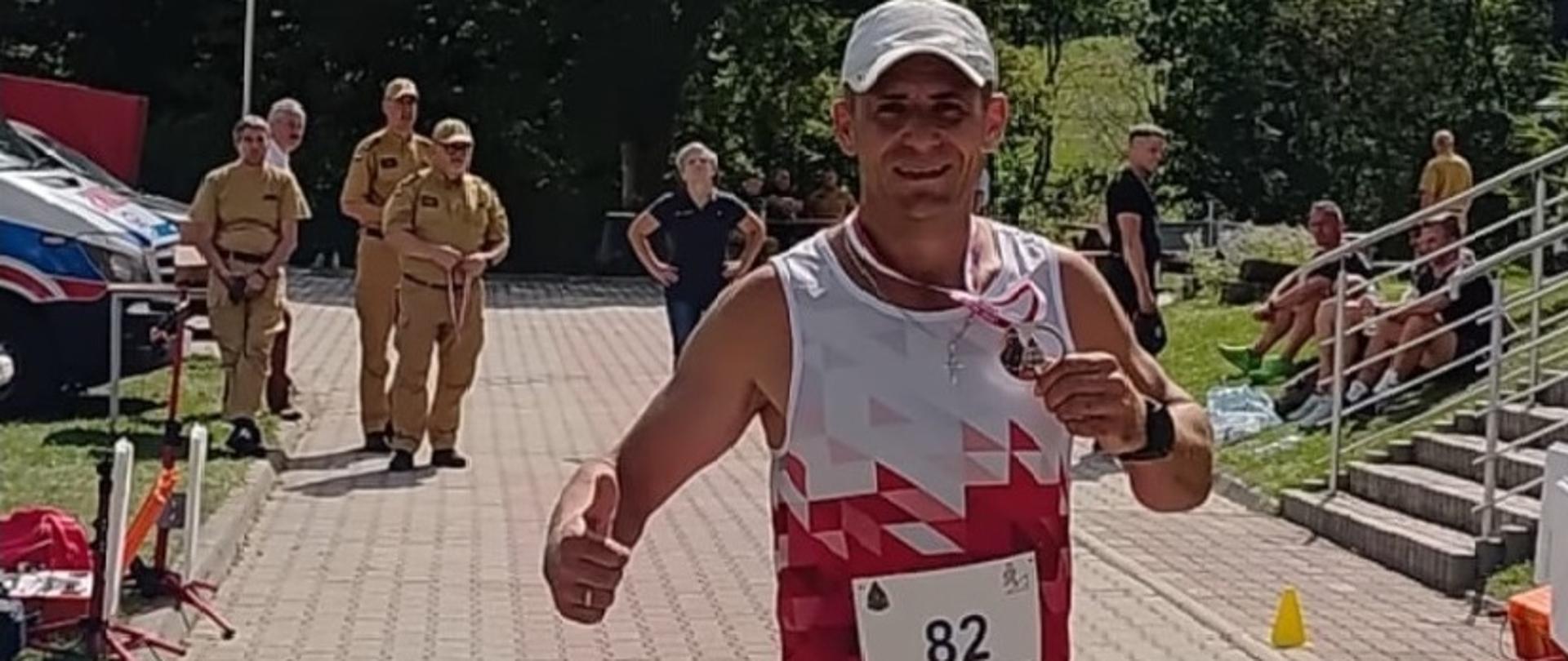 Kolorowa fotografia wykonana w pogodny dzień na zewnątrz. Przedstawia strażaka w stroju sportowym z czapką na głowie i numerem startowym na koszulce z wręczonym medalem po biegu w zawodach wojewódzkich.
W tle strażacy w umundurowaniu służbowym.

