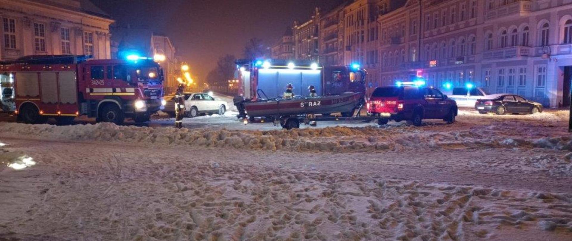 Na zdjęciu widać trzy samochody należące do Państwowej Straży Pożarnej, dwa ciężarowe i jeden osobowy, do którego podczepiona jest przyczepa z łodzią. Pojazdy mają włączoną sygnalizację ostrzegawczą. Na ich bocznej karoserii widnieją numery operacyjne. Z prawej strony zdjęcia można zobaczyć również radiowóz Policji, również z włączoną sygnalizacją ostrzegawczą. W tle widać zabudowania ulicy Aleja Wolności wraz z lampami oświetlenia ulicznego. Na miejscach parkingowych stoją zaparkowane samochody osobowe. Zdjęcie zostało wykonane w porze nocnej, na pierwszym planie widoczny jest śnieg zalegający na chodniku.