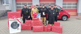 Strażacy stoją przed budynkiem komendy, z tyłu stoi samochód. Przed strażakami paczki z prezentami oraz pralka. 