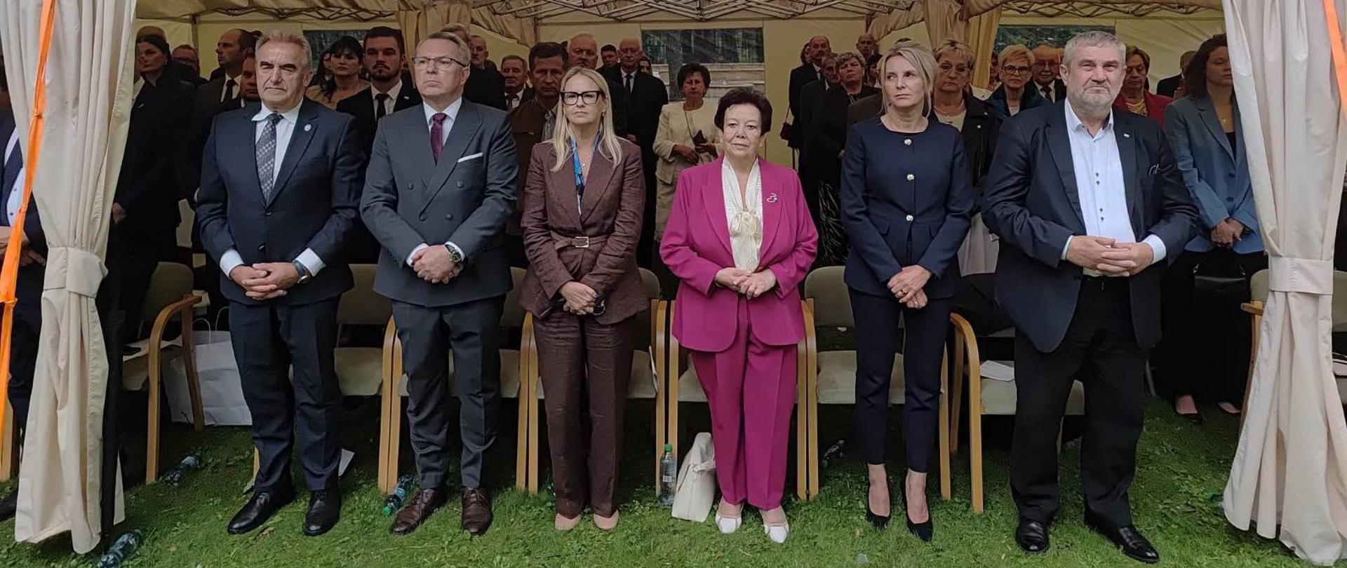 Grupa sześciu osób stoi w rzędzie pod dużym, białym namiotem bankietowym rozstawionym. Wszyscy są ubrani elegancko (garnitury, garsonki), patrzą w stronę obiektywu z powagą, z dłońmi splecionymi przed sobą. W tle widać zgromadzoną publiczność.