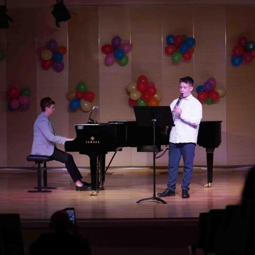 zdjęcie przedstawia ucznia grającego na klarnecie, przed nim pulpit, obok akompaniatorka gra na fortepianie, w tle dekoracja z kolorowych balonów