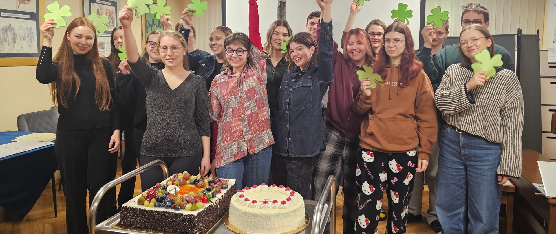 Na zdjęciu grupa młodzieży - 16 osób, które stoją razem, koło siebie. Wszyscy są uśmiechnięci, radośni. Kończą bowiem ważny etap życia - przed nimi matury i końcowe dyplomy. Wszyscy trzymają w rękach papierowe koniczynki jako znak szczęścia i pomyślności na przyszłość. Przed nimi stoi stolik na nim dwa duże torty - dla nich na osłodę i dodanie sił przed egzaminami