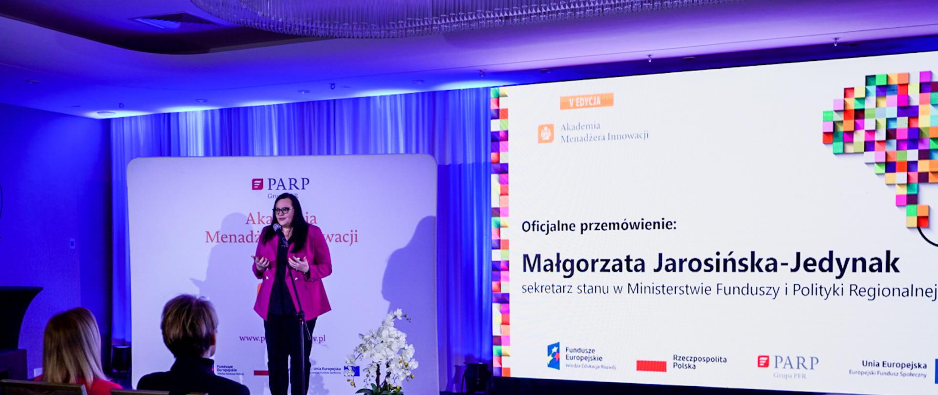 Wiceminister Małgorzata Jarosińska-Jedynak stoi na scenie i przemawia do ludzi zgromadzonych na sali.