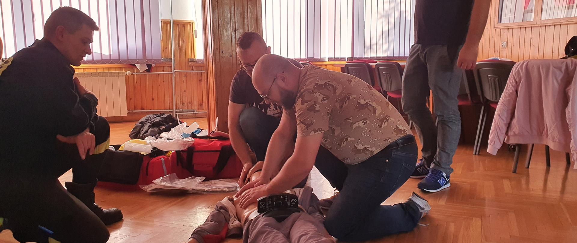 cztery osoby na zdjęciu, dwie osoby zajmują się prowadzeniem pierwszej pomocy na manekinie leżącym na podłodze, jeden funkcjonariusz nadzoruje ćwiczenie, jedna osoba to obserwator