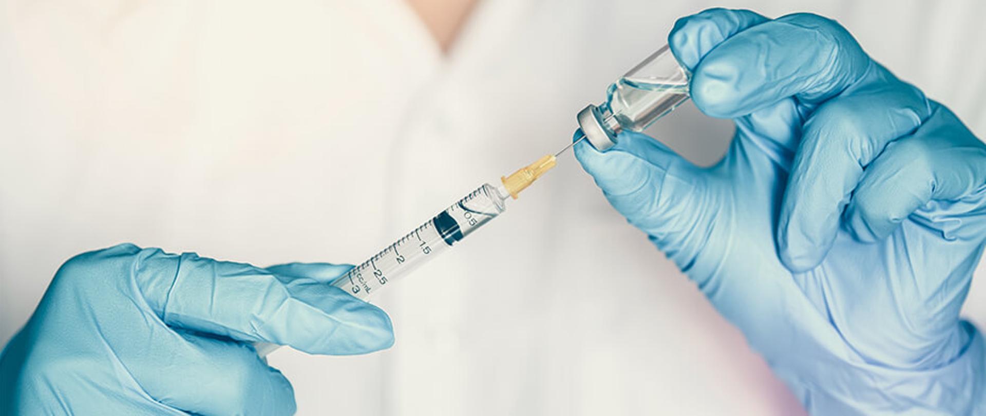 Zdjęcie, na którym widnieje fiolka ze szczepionką oraz strzykawka trzymane w dłoniach przez medyka.