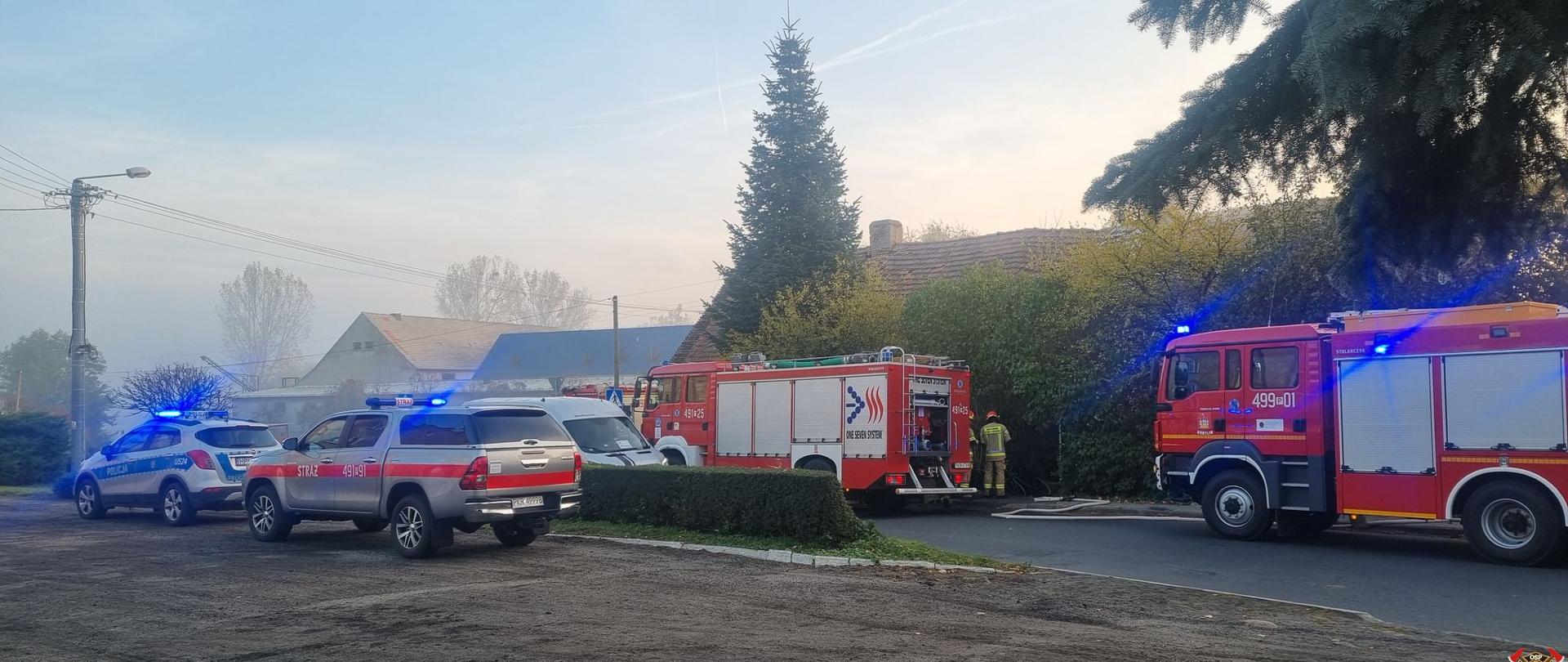 Na drodze przy opuszczonym budynku mieszkalnym stoją pojazdy pożarnicze z włączoną sygnalizacją świetlną. Obok pojazdu stoją strażacy obsługujący autopompy. Na chodniku widać przebiegające linie gaśnicze nawodnione. Na placu manewrowym przy drodze stoi radiowóz policyjny oraz pojazd rozpoznawczo ratowniczy. Trwa akcja gaśnicza
