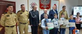 Kolorowa fotografia wykonana wewnątrz pomieszczenia. Przedstawia część sali z grupą stojących osób cywilnych i umundurowanymi strażakami. Przed nimi stoją młodzi uczestnicy turnieju z dyplomami i nagrodami. Na ścianach widoczne znaki i symbole strażackie oraz gabloty z pucharami, stojący baner informacyjny, panel wyświetlacza, Godło Polski i dyplomy. 