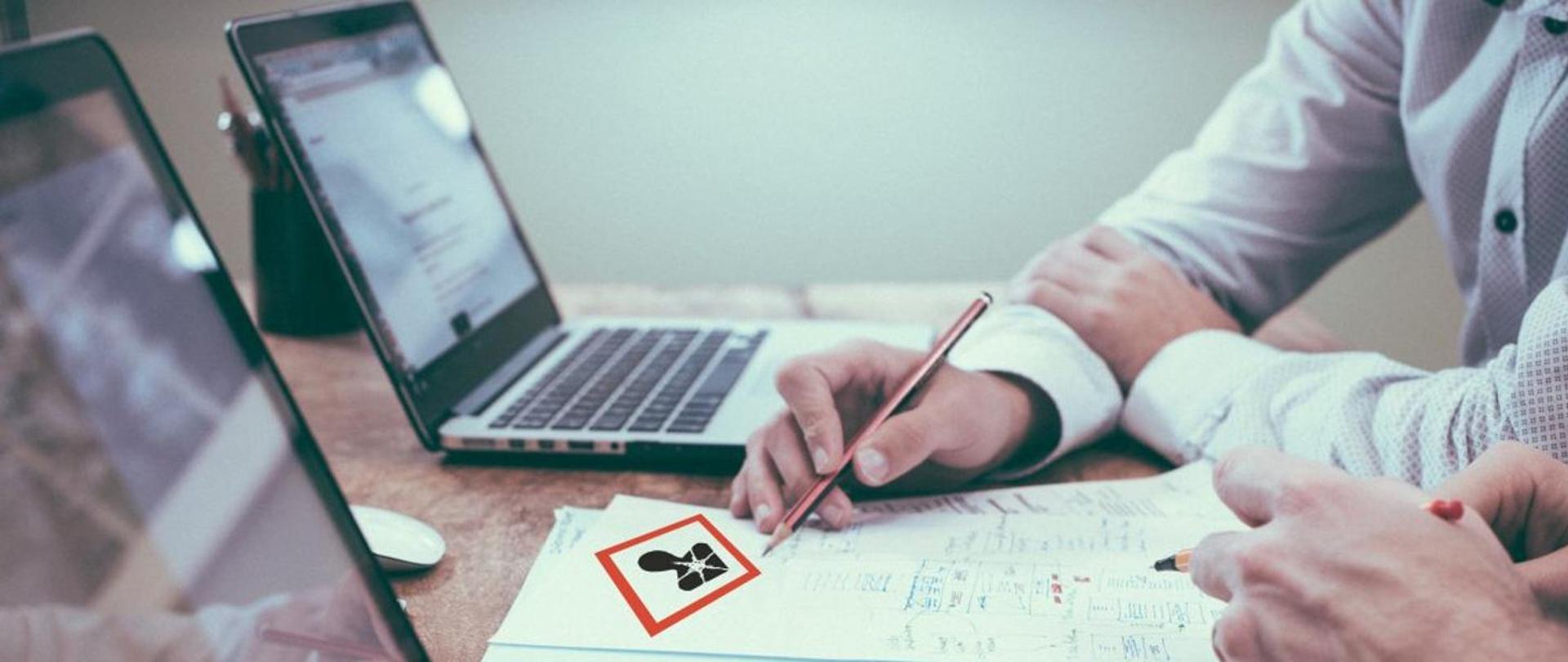Zdjęcie przedstawia dwa otwarte laptopy oraz dłonie mężczyzn z długopisami, którzy notują informacje na kartkach. Na środku piktogram dla oznakowania działania rakotwórczego