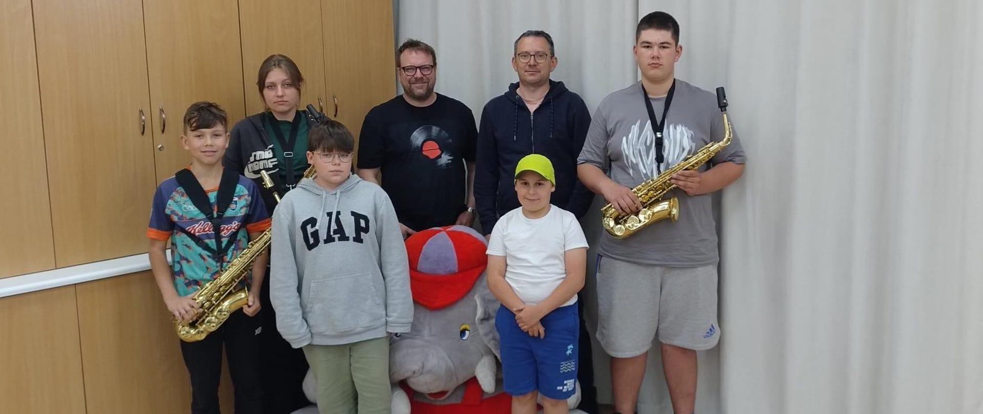 Grupa osób stoi w klasie i pozuje do zdjęcia. Wśród nich jest czterech chłopców, jedna dziewczynka i dwóch mężczyzn. Dwie osoby trzymają w ręku saksofony. Pomiędzy dwoma chłopcami na podłodze stoi duża maskotka przedstawiająca słonia.