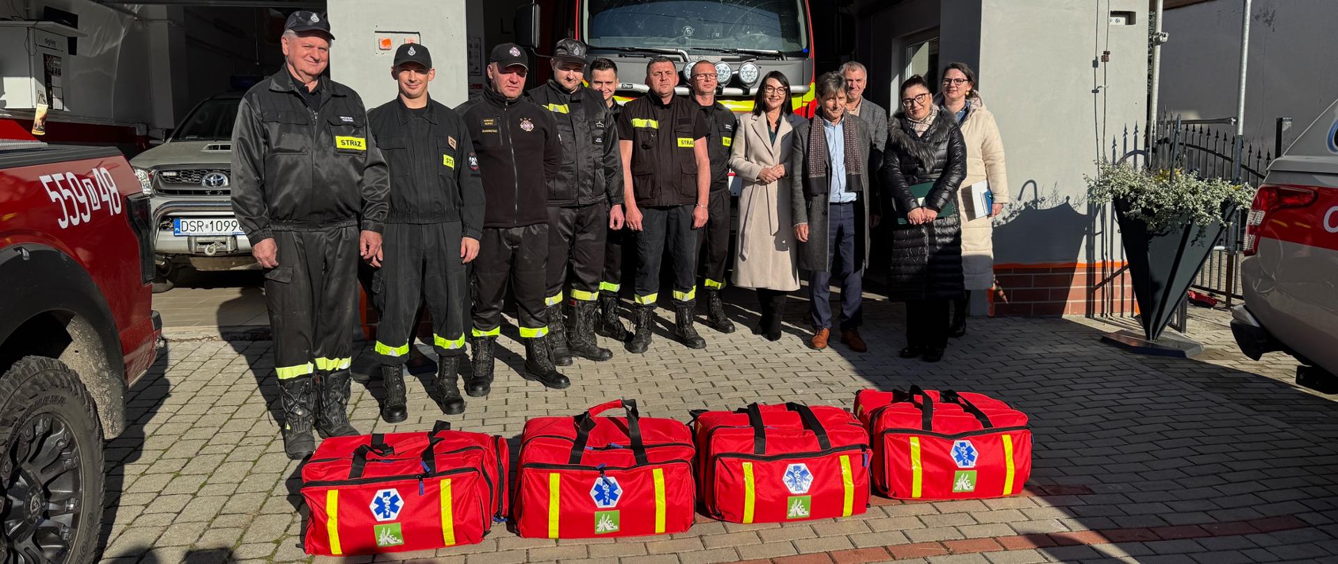 Przed wozem strażackim z napisem „OSP Brodno” ustawiona jest grupa strażaków w mundurach oraz kilka osób cywilnych. Wszyscy stoją w rzędzie na tle remizy. Na ziemi przed nimi ułożonych jest sześć czerwonych toreb ratowniczych z niebieskimi symbolami medycznymi.