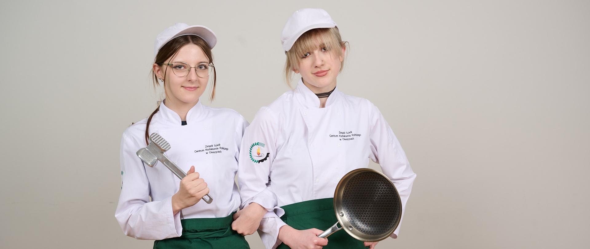 Dwie uczennice stoją w strojach kucharskich: biała bluza z logo szkoły i czapka, zielona zapaska. W prawej ręce jedna trzyma tłuczek do mięsa , a druga patelnię. Lewą rękę trzymają opartą na biodrze.