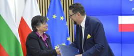 Przy stoliku stoją wiceminister Szeptycki i kobieta w czarnej marynarce, podają sobie ręce,, za nimi pod ścianą flagi Polski, Bułgarii i UE.