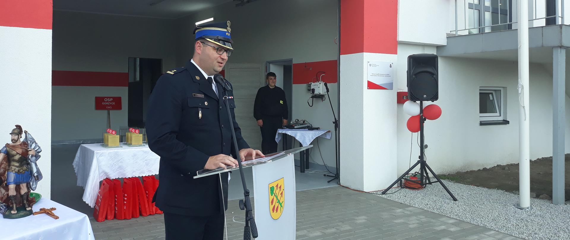 strażak w ciemnym mundurze i w rogatywce stoi przy mównicy, w tle statuetka świetego floriana