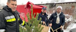 Trzej mężczyźni - Strażak trzyma w ręce choinkę, Policjant trzyma torbę z ozdobami świątecznymi, i pracownik MOPS trzyma stojak do choinki i walizkę. W tle strażacka przyczepka oraz budynek mieszkalny wielorodzinny.