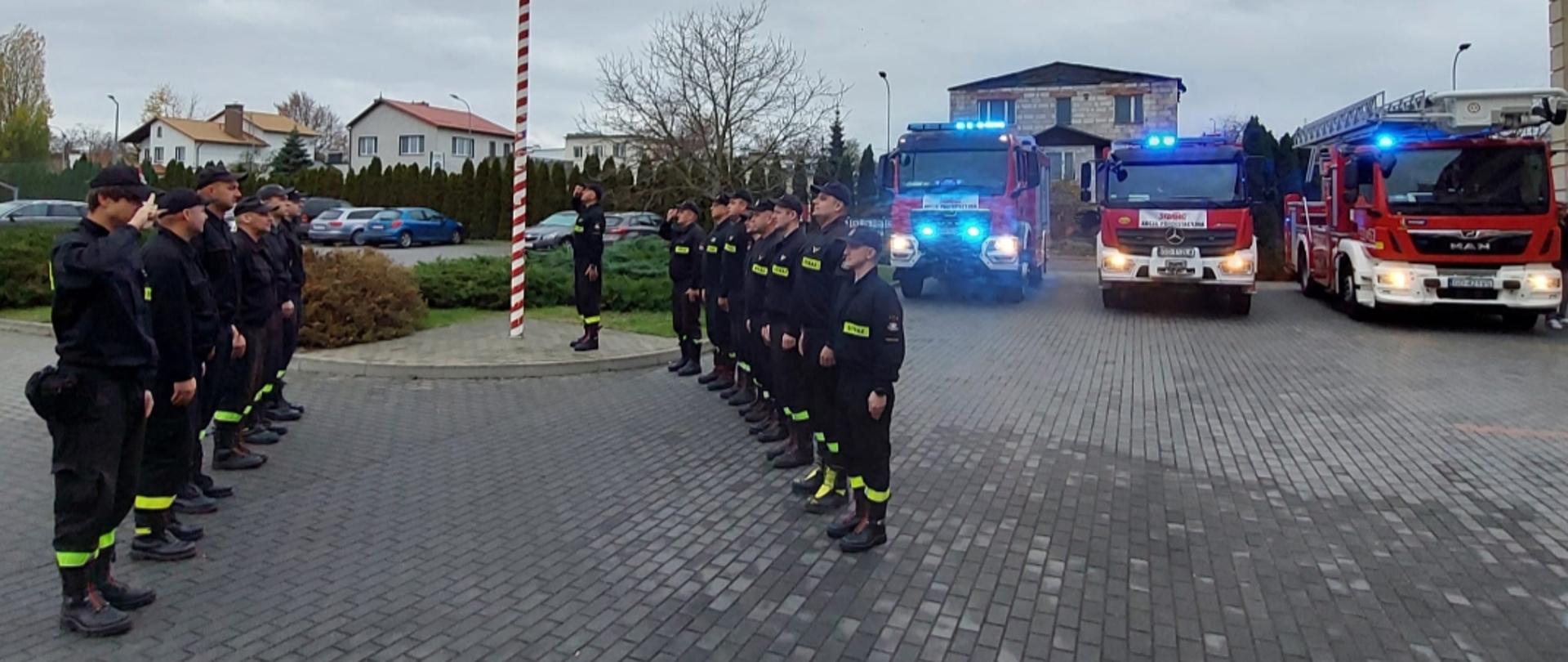 Zdjęcie przedstawia strażaków podczas uroczystego podnoszenia flagi na maszt, w tle widać samochody strażackie.
