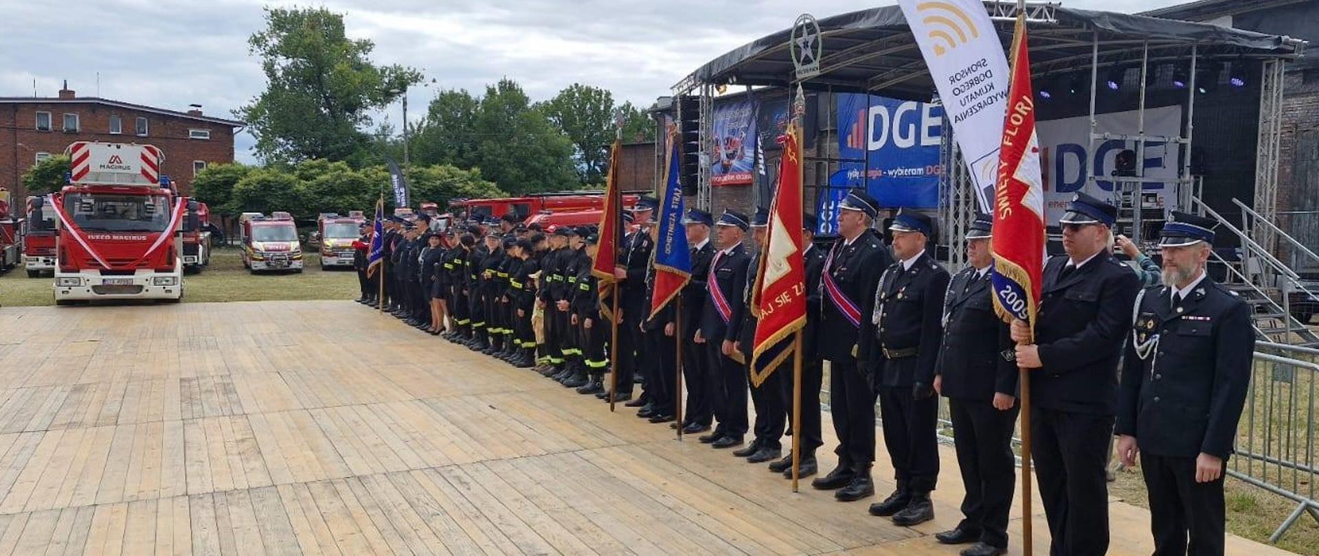Drewniany podest. Na nim w rzędzie stoją strażacy. Niektórzy trzymają sztandary. Na wprost czerwony samochód strażacki.