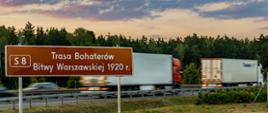 Zdjęcie przedstawiające tablicę informującą o poruszaniu się drogą ekspresową S8, Trasą Bohaterów Bitwy Warszawskiej 1920 r. W tle widać poruszające się drogą ekspresową pojazdy