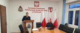 Na zdjęciu przy mównicy na sali konferencyjnej referuje komendant powiatowy PSP w Gorlicach w mundurze wyjściowym granatowym obok niego przy stole siedzi oficer w ubraniu czarnym dowódczo sztabowym za nim trzy biało czerwone flagi.