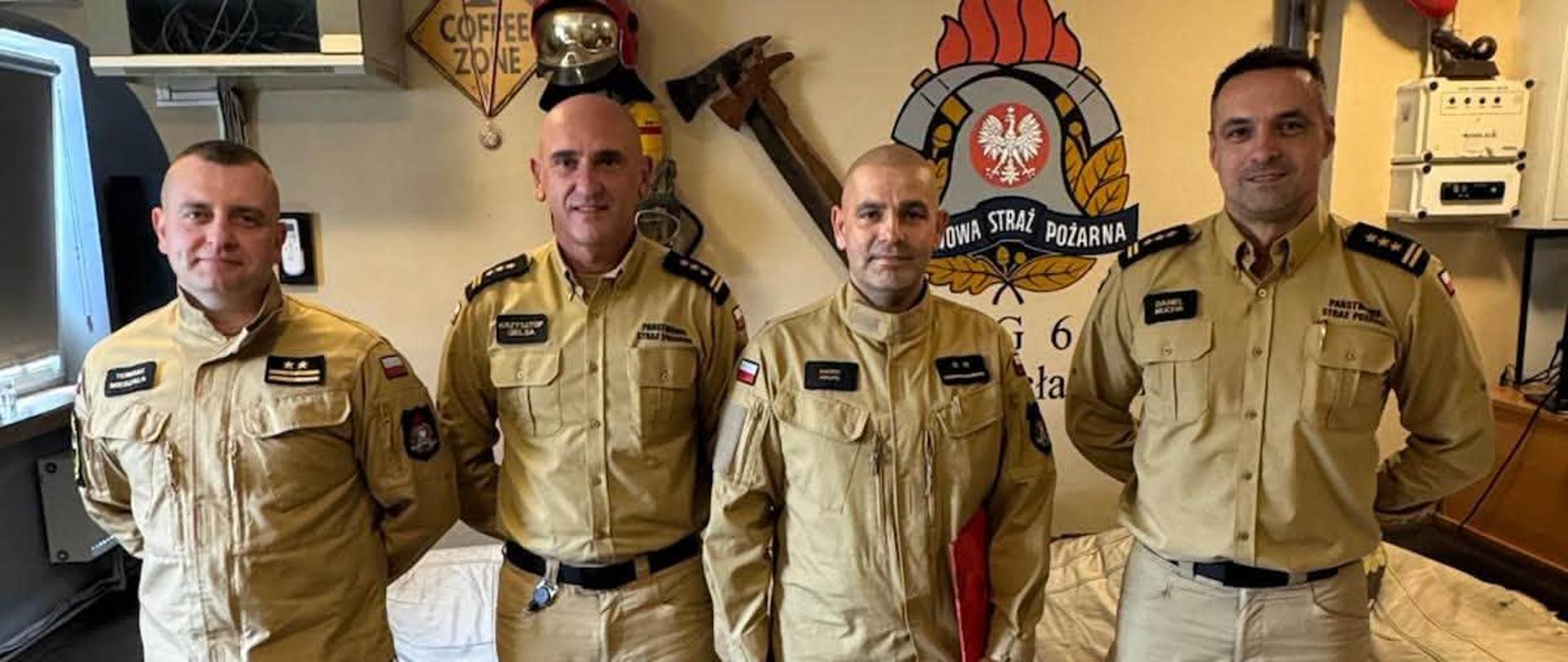Czterech strażaków Państwowej Straży Pożarnej w mundurach służbowych pozuje do wspólnego zdjęcia w pomieszczeniu. W tle widoczne są emblemat PSP, elementy wyposażenia strażackiego oraz sprzęt techniczny.