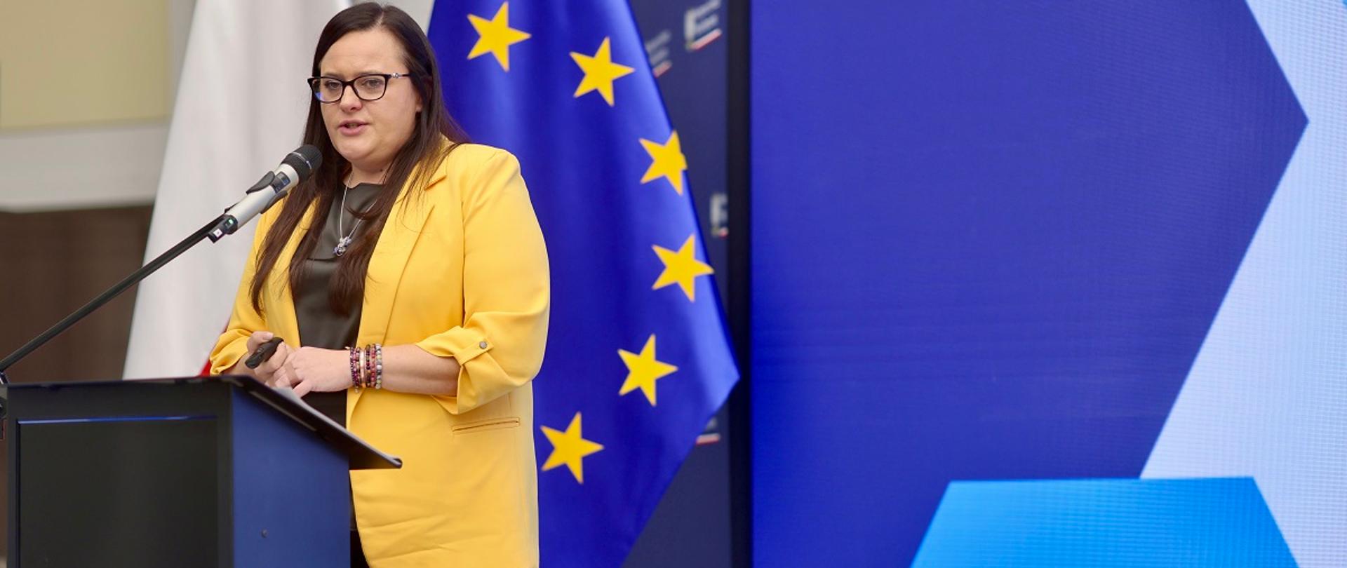 Minister funduszy i polityki regionalnej Małgorzata Jarosińska-Jedynak stoi przy pulpicie i przemawia do mikrofonu, za jej plecami flagi Polski i UE