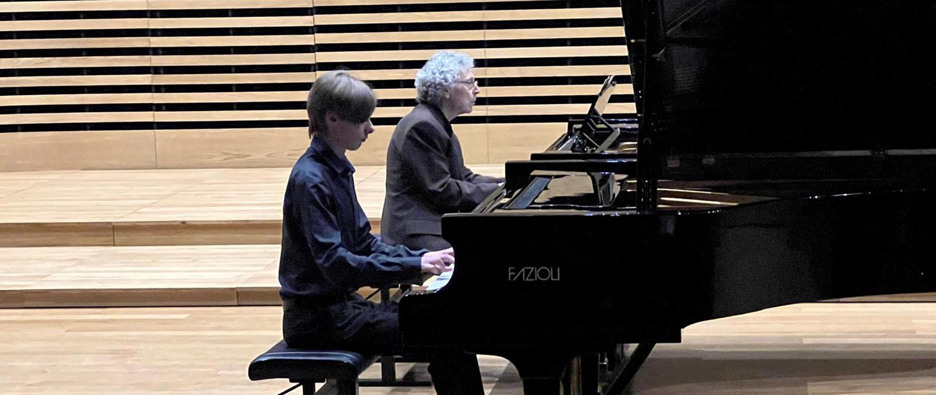 Chłopak i mężczyzna grają na dwóch fortepianach.