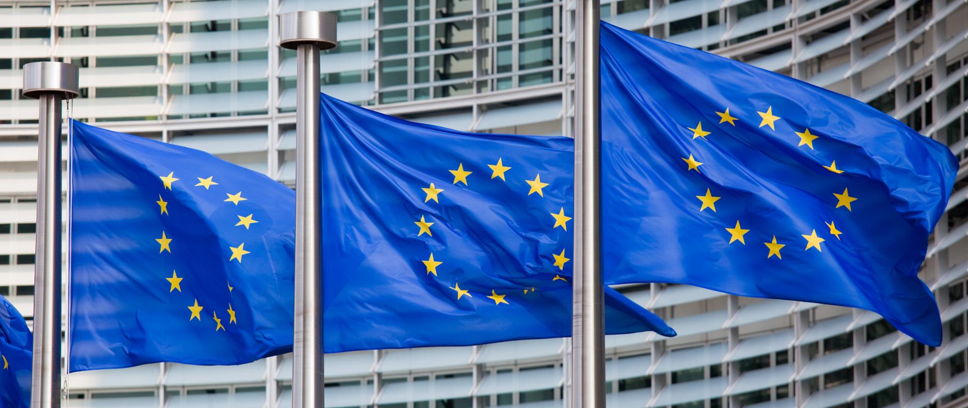 Trzy maszty z flagami Unii Europejskiej stojące na tle gmachu Parlamentu Europejskiego