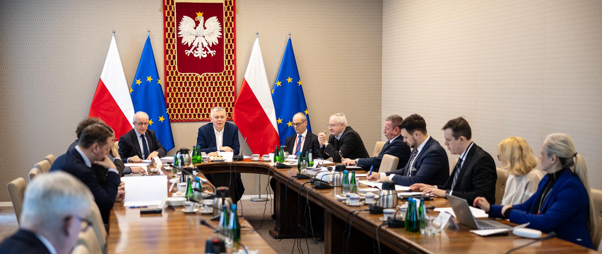 Grupa osób siedzących przy stole. W tle flagi polskie i europejskie oraz godło Polski 