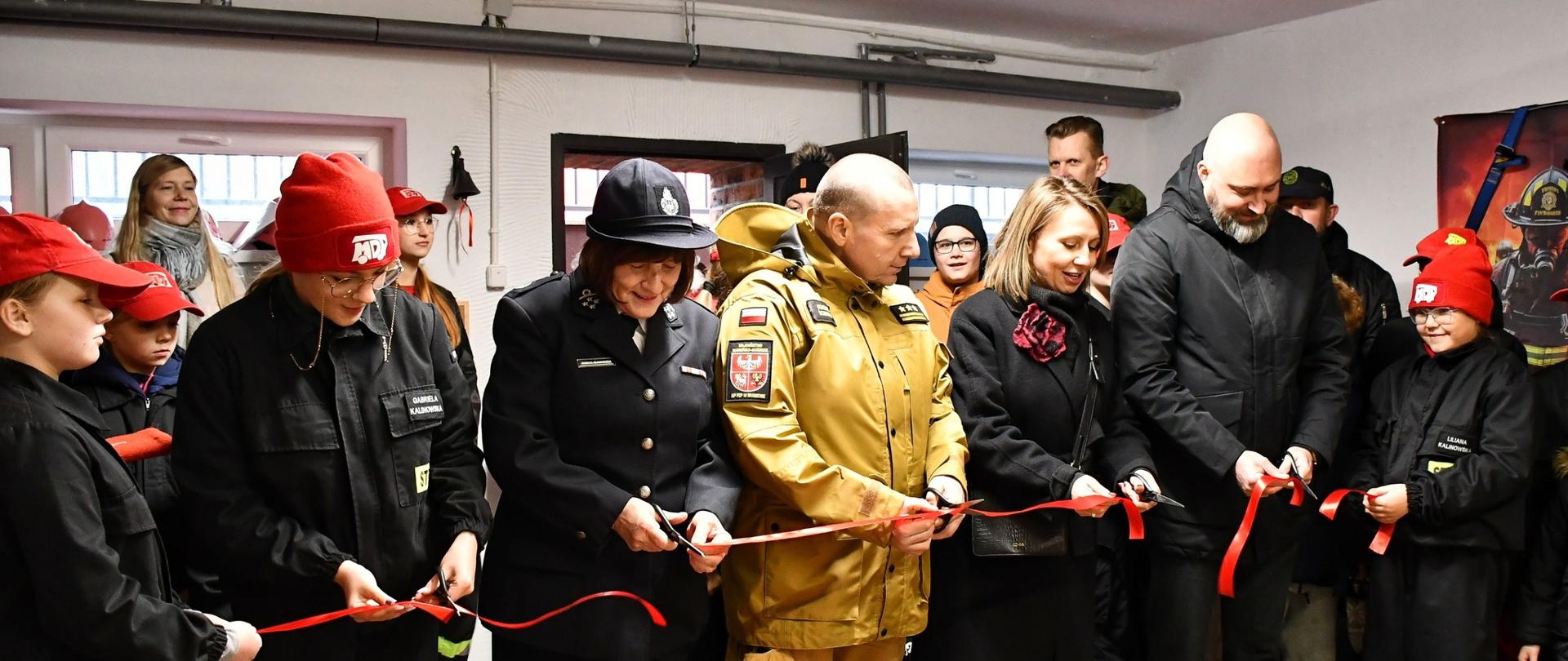 Trwa przecinanie wstęgi nowo otwartej salki edukacyjnej. Na zdjęciu strażacy, dzieci i samorządowcy.