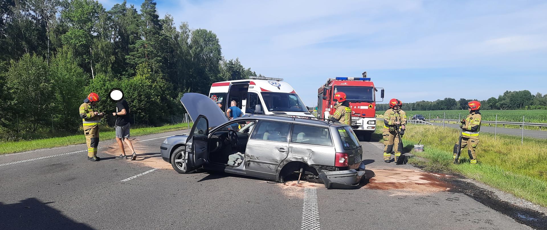 Na jezdni ustawiony w poprzek uszkodzony szary samochód osobowy. Przy aucie strażacy sprzątają wycieki płynów. Za nimi karetka i samochód strażacki.