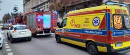 Na zdjęciu widzimy karetkę pogotowia koloru żółtego i czerwonego. Karetka stoi na ulicy przed budynkami mieszkalnymi. Dalej widzimy samochody strażackie koloru czerwonego. Obok samochodów przejeżdżają samochody osobowe. Dalej w tle widzimy budynki wielorodzinne.