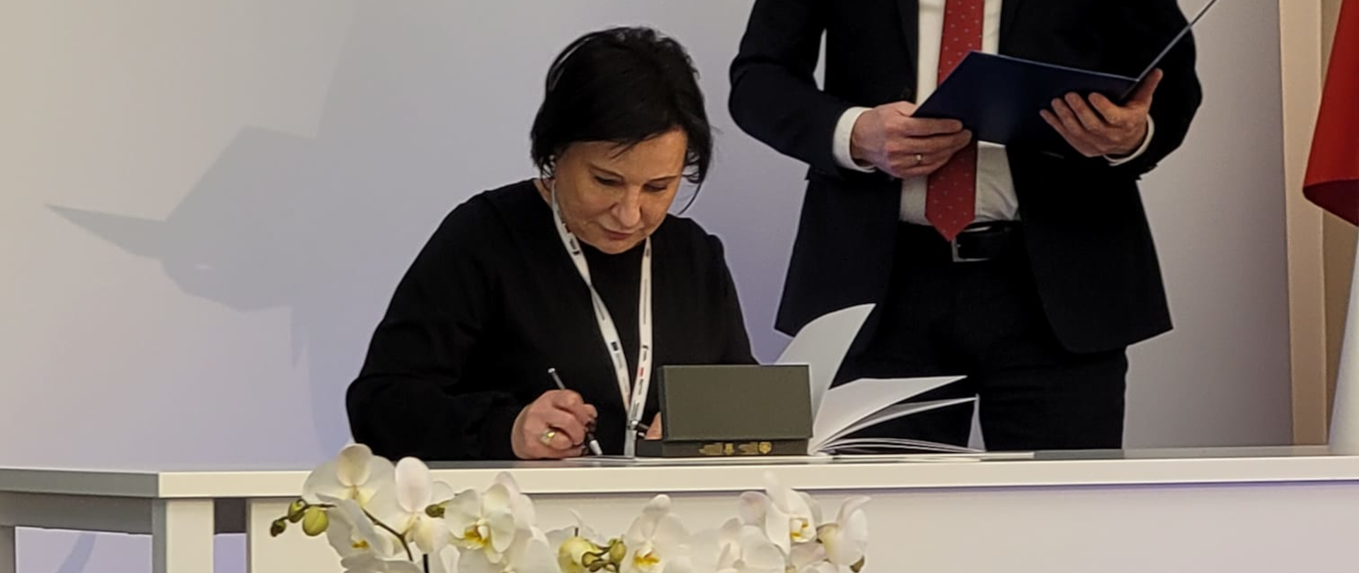 na tle białego banera z napisem Prosto i kropka siedzi przy biurko kobieta i podpisuje dokumenty, obok stoi mężczyzna w garniturze i trzyma w dłoniach otwarty dyplom w twardych okładkach