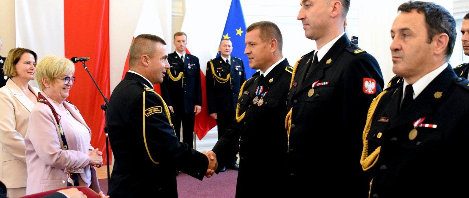 Zdjęcie zrobione w sali kolumnowej podkarpackiego Urzędu Wojewódzkiego podczas uroczystego apelu. Oficer Państwowej Straży Pożarnej w stopniu starszego brygadiera gratuluje oznaczenia strażakowi ochotniczej straży pożarnej.