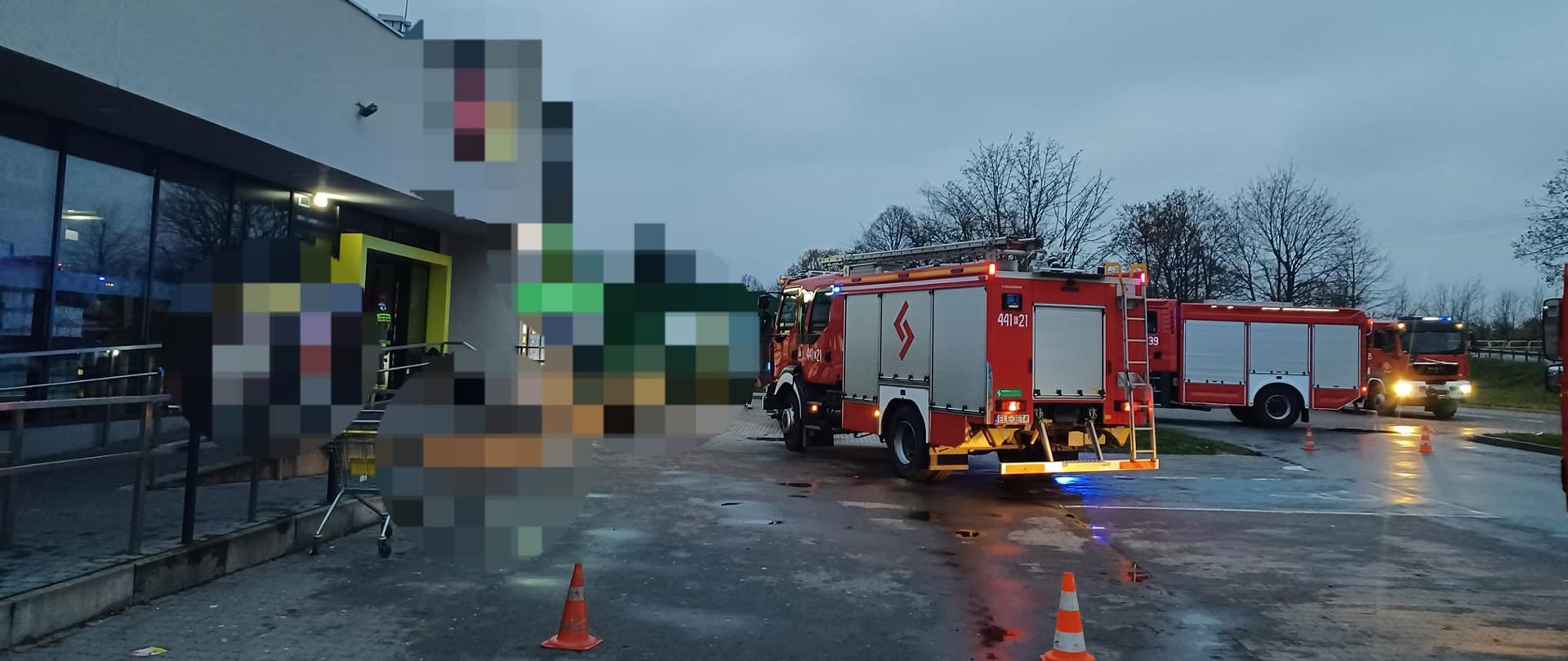 Na zdjęciu widać trzy czerwone samochody strażackie stojące na parkingu supermarketu. Jeden strażak w czerwonym hełmie i żółtym stroju stoi przed wejściem. Na parkingu ustawione są pomarańczowo-białe pachołki. W tle widać drzewa. Jest widno, ale pochmurnie.
