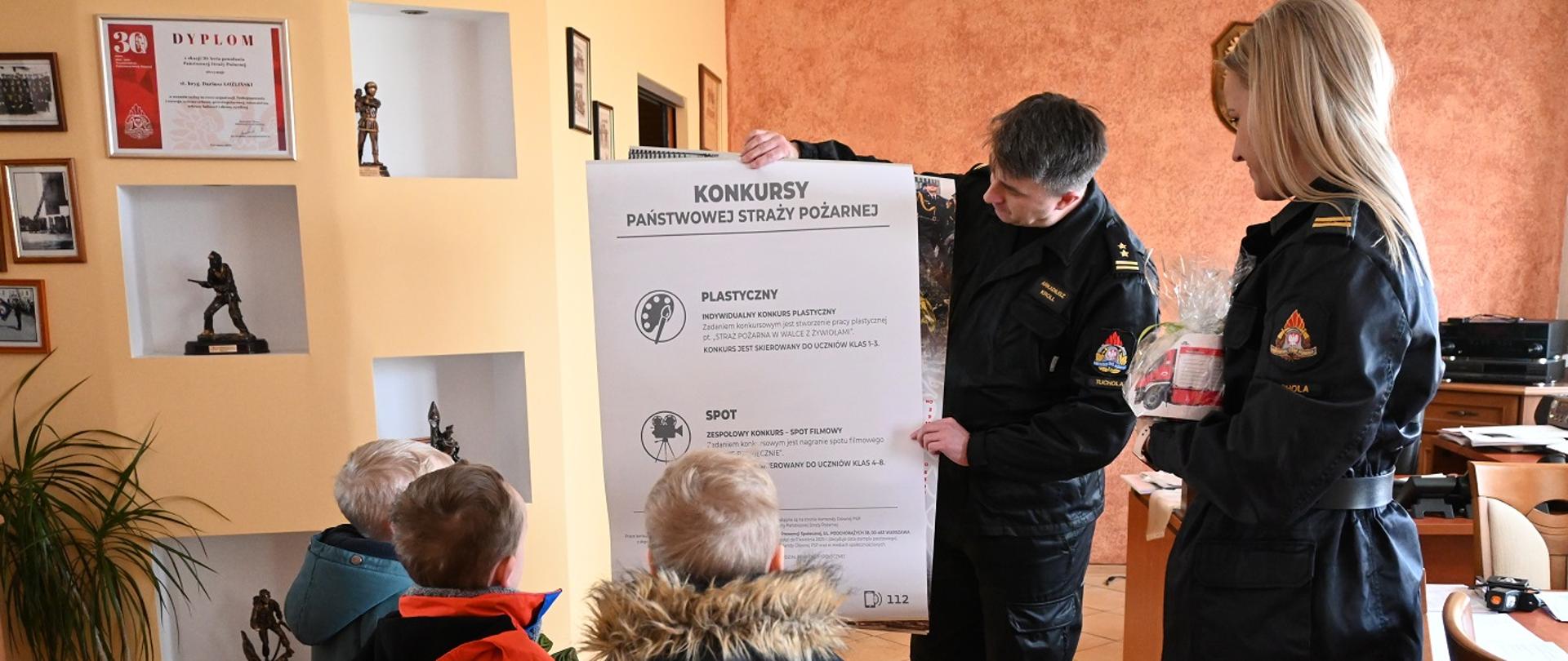 Na zdjęciu strażak w czarnym mundurze dowódczo-sztabowym prezentuje kalendarz Państwowej Straży Pożarnej w ogłoszonymi konkursami. Zachęca dzieci do wzięcia udziału. Obok niego stoi funkcjonariuszka straży pożarnej w tym samym mundurze, a naprzeciwko stoi 3 chłopców.